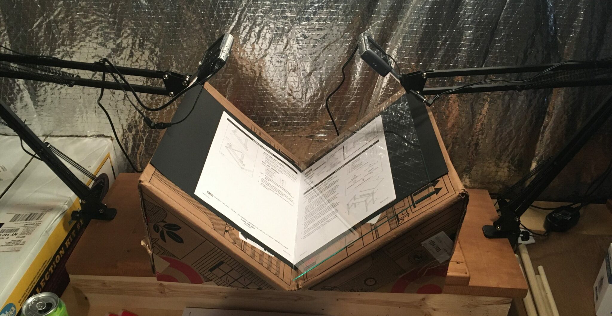 A close-up photograph of the book scanning rig, which is currently a cardboard box cut into the shape of a lectern, and two digital photography cameras on articulated arms, each pointed at one page of the open book, as it sits on the rig.