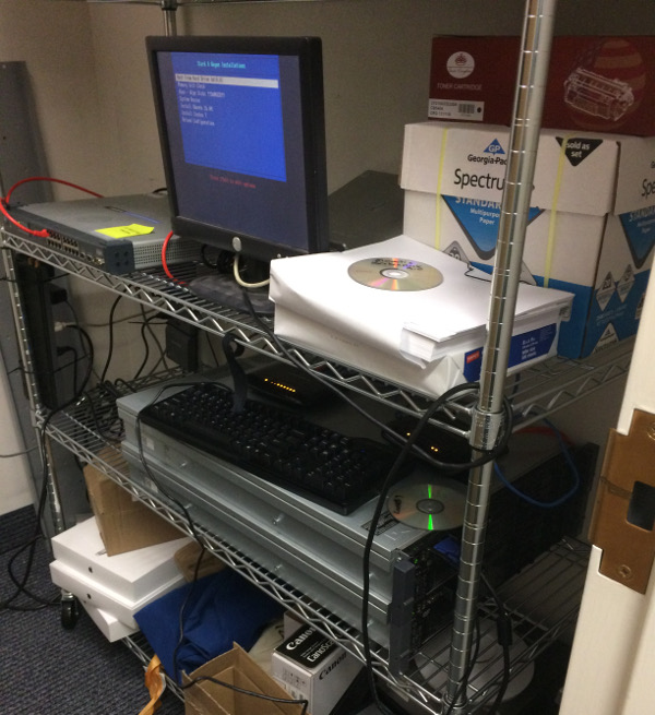 Picture of the wiring closet where we keep the lab servers, while we operate on them.  A monitor sits on a wire shelf, with two rack-mount Dell 2U servers stacked one on top of the other on the shelf below.  The monitor is displaying a PXE boot screen.