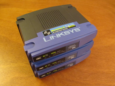 Photograph of a stack of 3 Linksys WRT 54G wireless router devices.